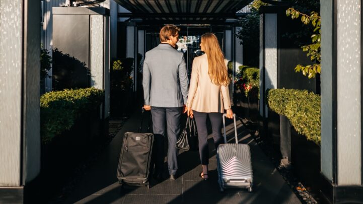 couple dressed corporate looking at each other while walking into a hotel resort and dragging their luggages behind them