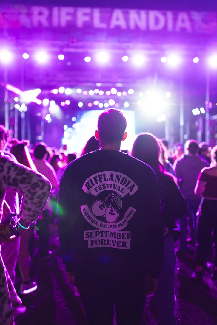 a crowd of people at a colorful lit stage event with tons of white and purple lights and a guy wearing a dark shirt tha reads "Rifflandia Festival, September Forever"