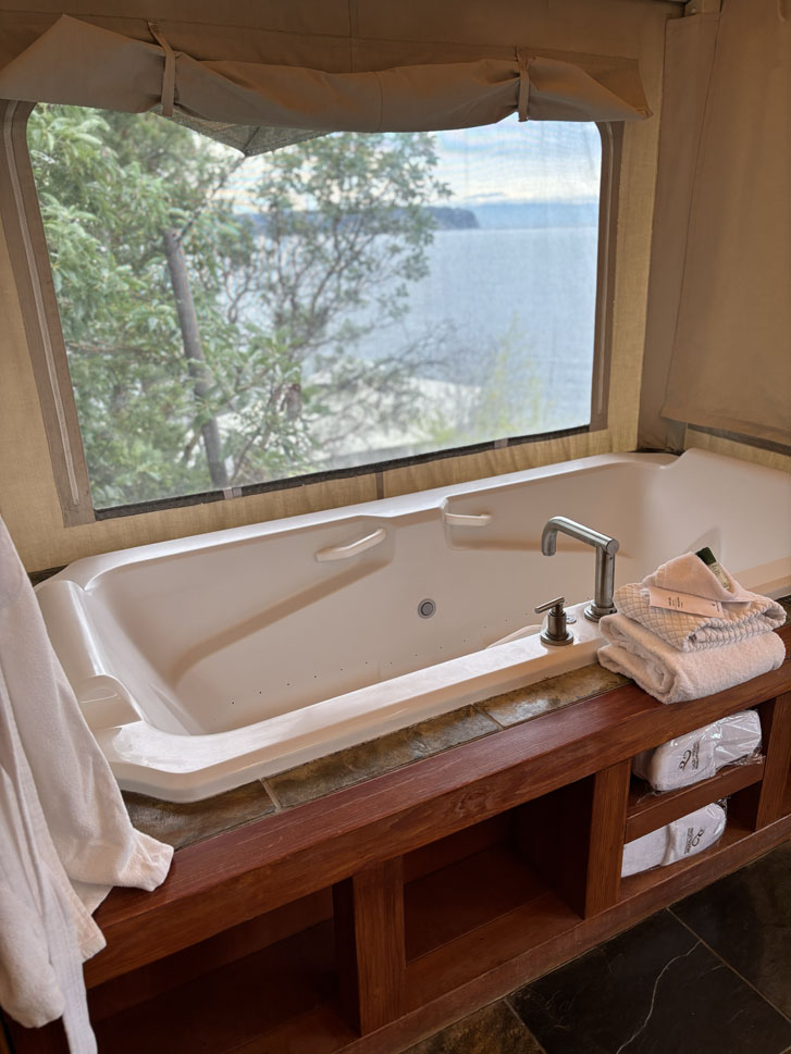 two-person soaker tub with white towels on the wooden side and a transparent window overlooking the trees and ocean