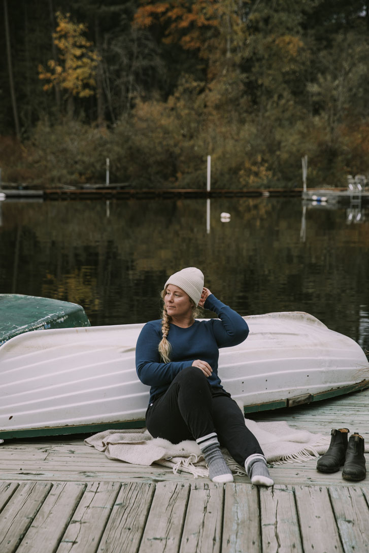 woman in covered fashion clothing, beanie, and socks, sitting on a deck with her black boots beside her and a turned-over boat behind her