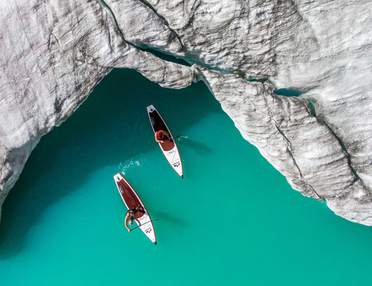 two paddlers paddling on a paddle board coming out of a rocky covering on a blue sea