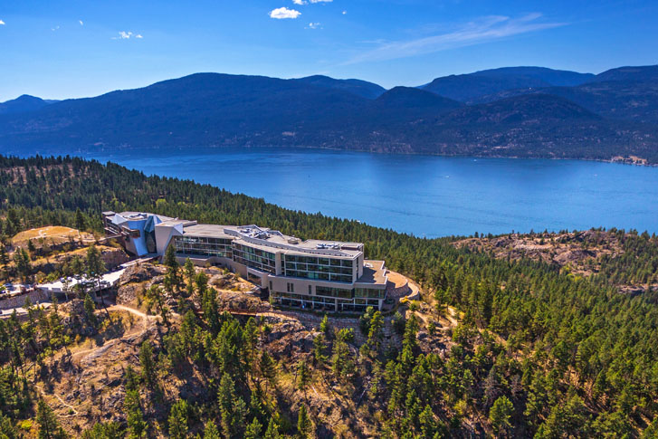 aerial view of a massive vast glass-windowed building in the middle of a foresty area overlooking the ocean and hills on the far end