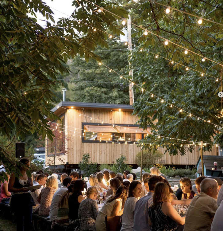 groups of people all seated on chairs outside a restaurant with little light bulbs across the trees above them