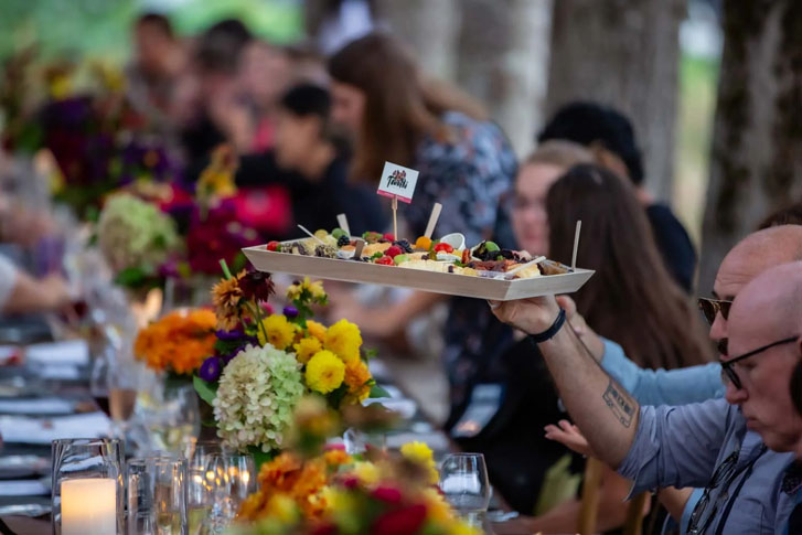 a group of people seated around an outdoor dinner table with a man passing a tray of tiny sweet fruits