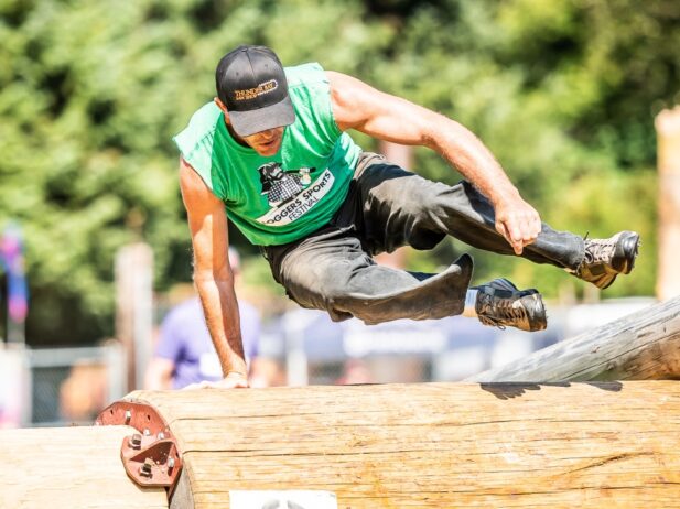 a man in a green sleeveless shirt and trousers jumping over a fat long log of wood that has the number sign "3" on it