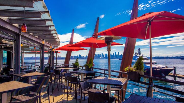 a patio restaurant with chairs, tables and red canopies around the patio and a huge river beside it with a white yacht or boat