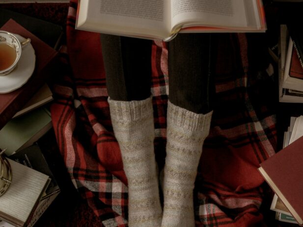 lady legs in socks with an open book on top, books laying on the floor and a cup of tea nearby