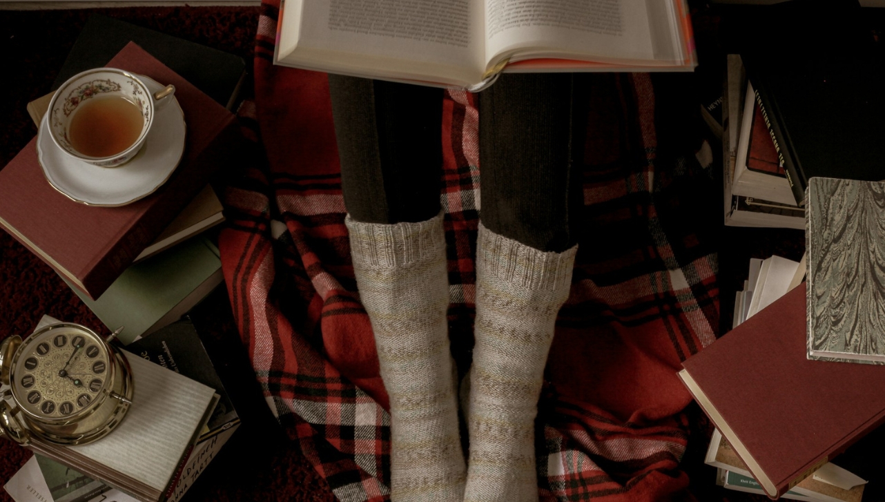 lady legs in socks with an open book on top, books laying on the floor and a cup of tea nearby