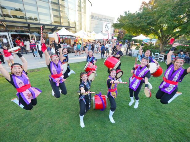 a group of females dressed in cultural attire, holding drums, drumsticks, and tambourines while jumping up in joy on a grass carpet field