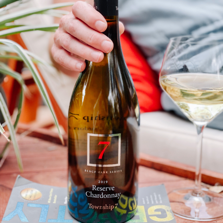 person holding the neck of a Township 7 chardonnay bottle with a glass with white wine next to it