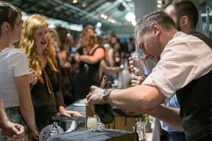 a man bartender poring some drink shots into tiny glasses for two ladies at a festival