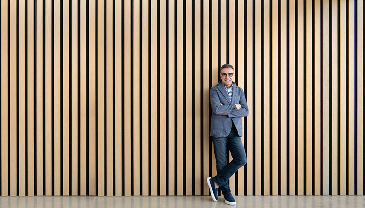 a smiling man leaning and standing in front of a wooden patterned wall with folded arms and legs