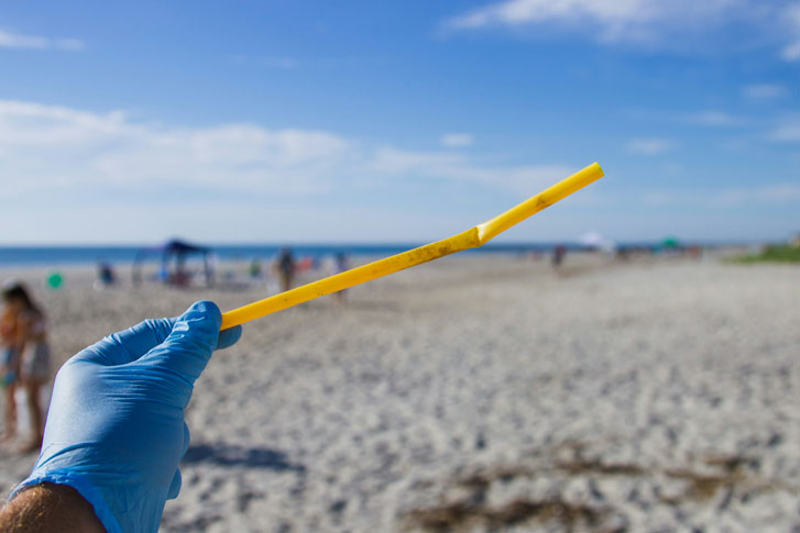 a left hand in blue glove holding up a sandy straw at a beach