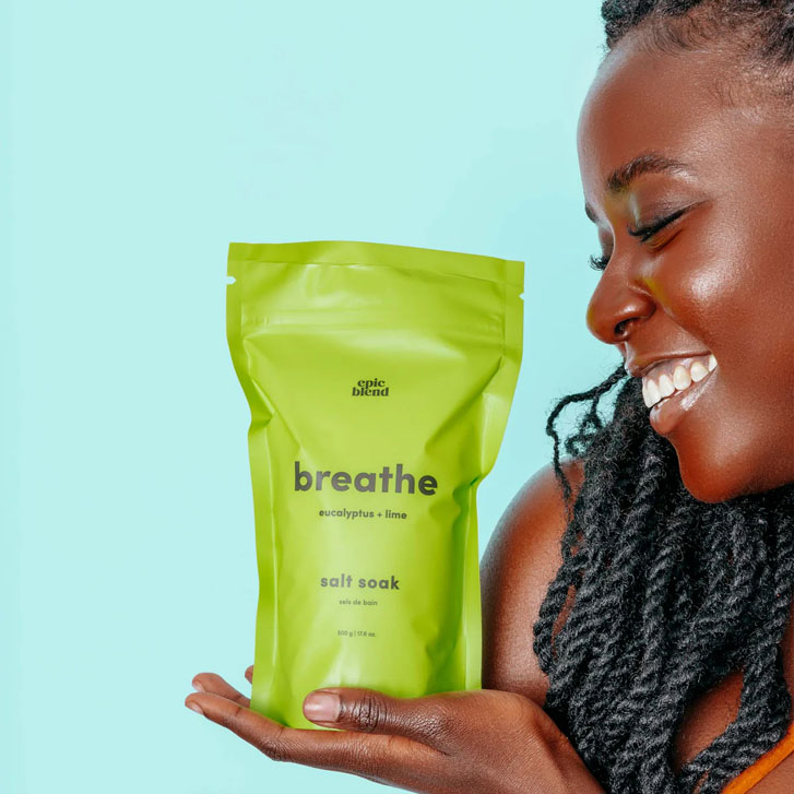 smiling black woman in braids holding a spa product on her hand that says"breathe, eucalyptus + lime Salt Soak"