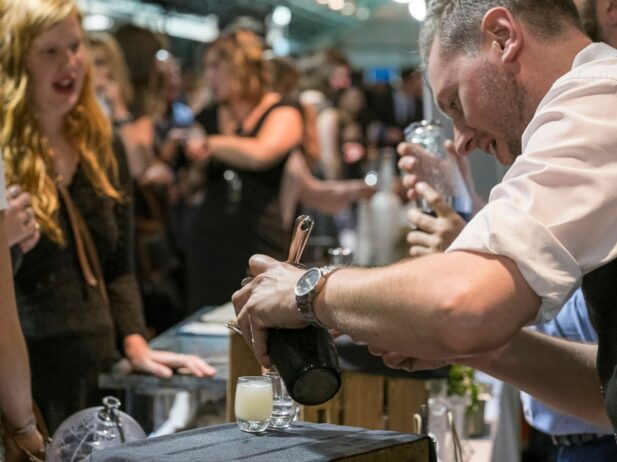 a man bartender poring some drink shots into tiny glasses for two ladies at a festival