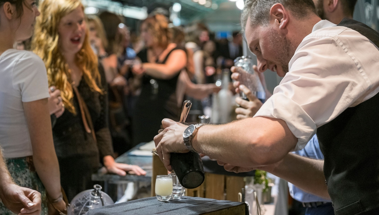 a man bartender poring some drink shots into tiny glasses for two ladies at a festival