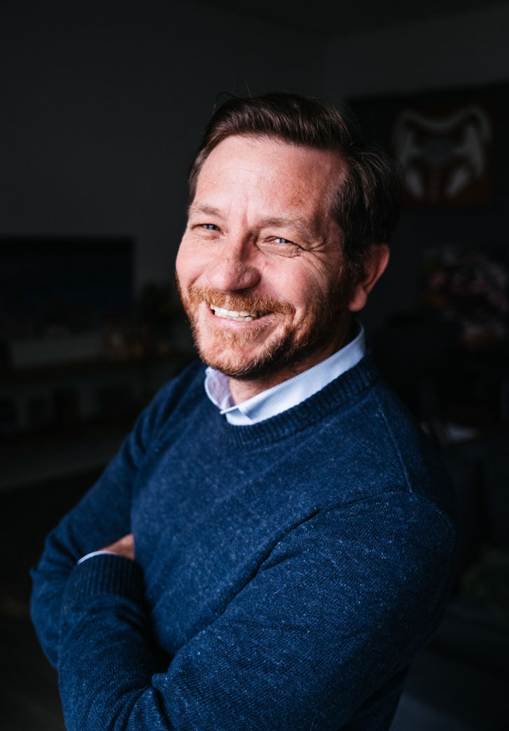 a smiling man in a professional shirt and sweater with his arms folded to the side