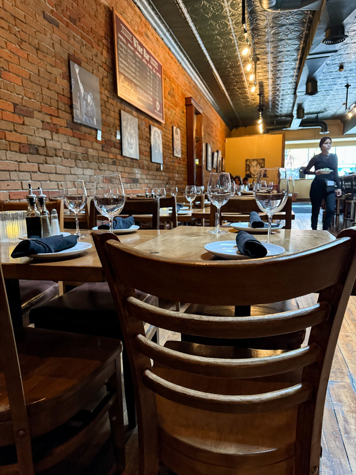 a girl walking past with a plate of food in a restaurant that has sets of glasses on the tables with chairs around