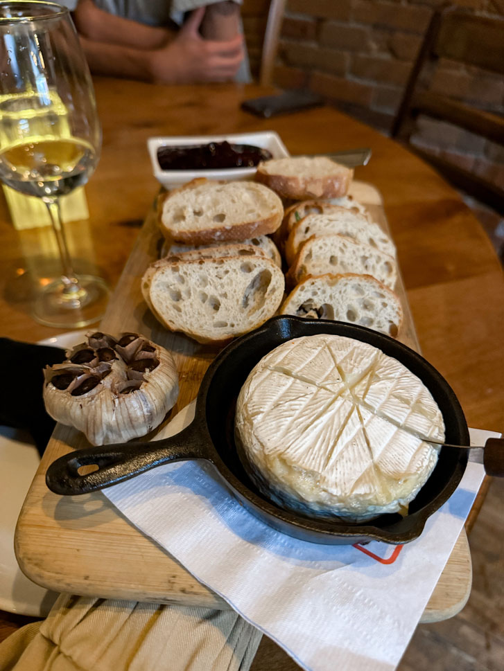 a wooden table with a glass of wine, slices of bread, a sauce and a pan of tiramisu