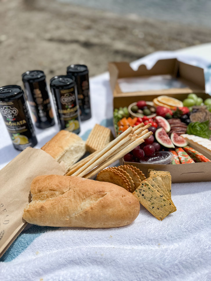 a super fresh charcuterie box with cheese, grapes, tomatoes, strawberries, ham and other veggies with breads and drinks beside