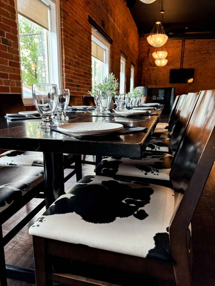 a brick themed room with a long table having chairs around it and plates and glasses on it with hanging lights above