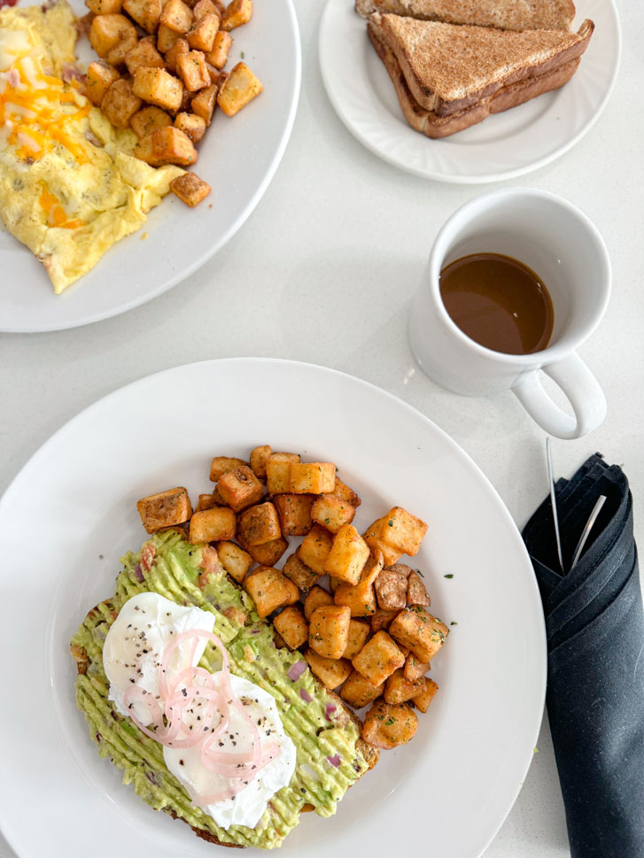 three white breakfast glass plates with avocado toast, eggs, potato cubes and a cup of tea