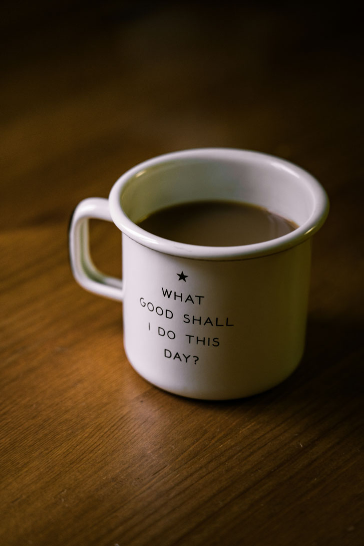 a cup of coffee that reads "What good shall I do this day" resting on a wooden table