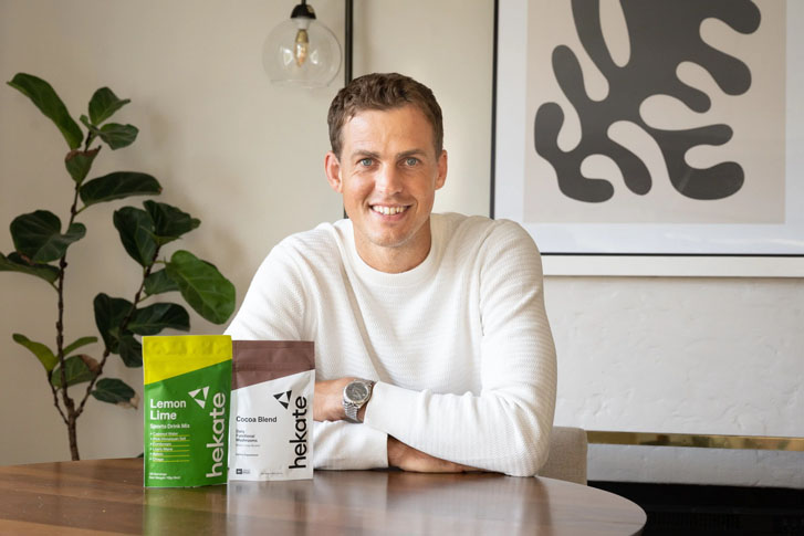 smiling man sitting in a room with his folded arms resting on a table that has two products that read "hekate"