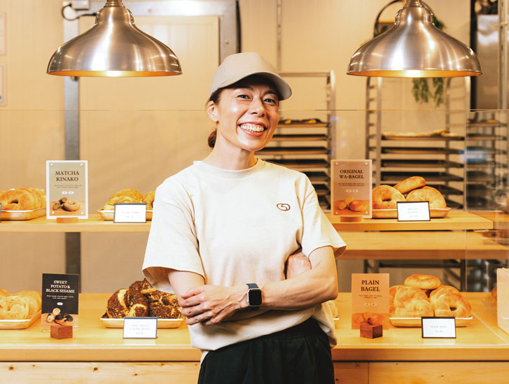 smiling Japanese woman standing in front of a food startup restaurant stand with groups of bagels and pastries behind her on the counter surfaces