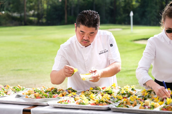 a chef in white placing vegetables on trays of food